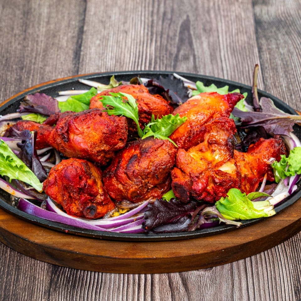 Sizzling tandoori chicken drumsticks with red onions and lettuce on a black platter.