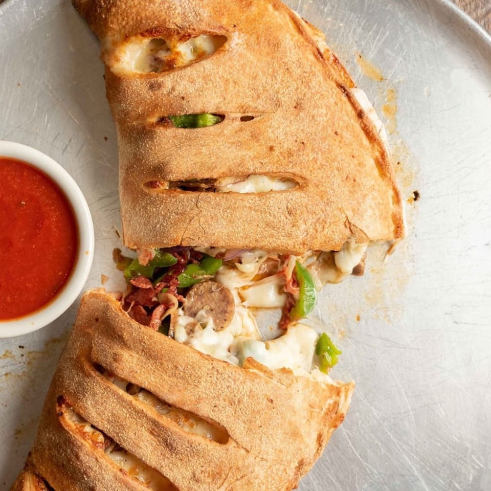 A Ricco's Pizza calzone, sliced open to reveal sausage, peppers, and melted cheese, with a side of marinara.