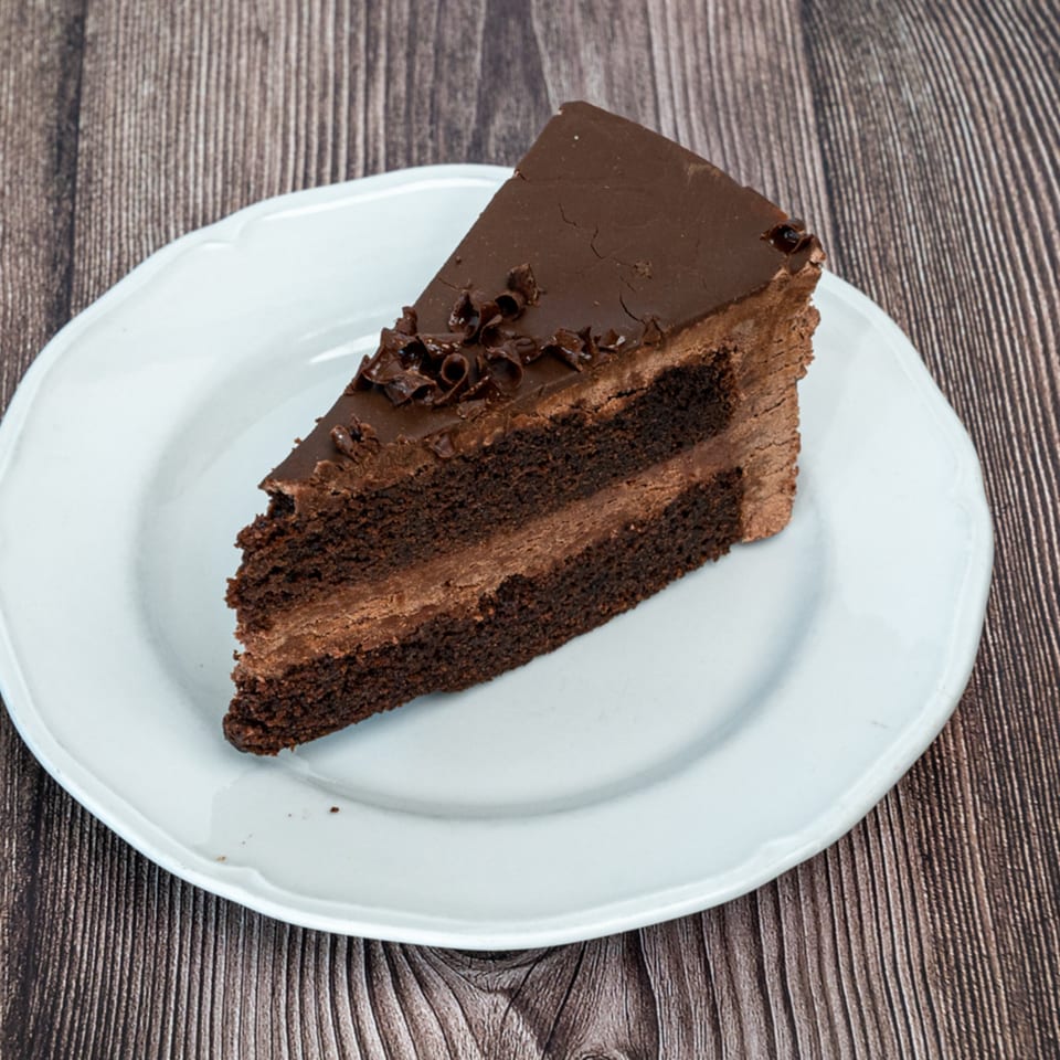 A slice of rich chocolate cake with chocolate frosting and shavings on a white plate.